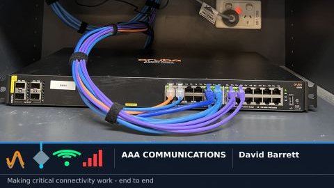 Aruba switch sitting outside a wall-mounted comms cabinet during a refresh, patched back into the rack with exposed leads