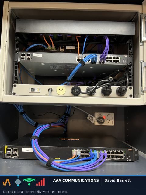 Aruba switch sitting outside a wall-mounted comms cabinet during a refresh, patched back into the rack with exposed leads