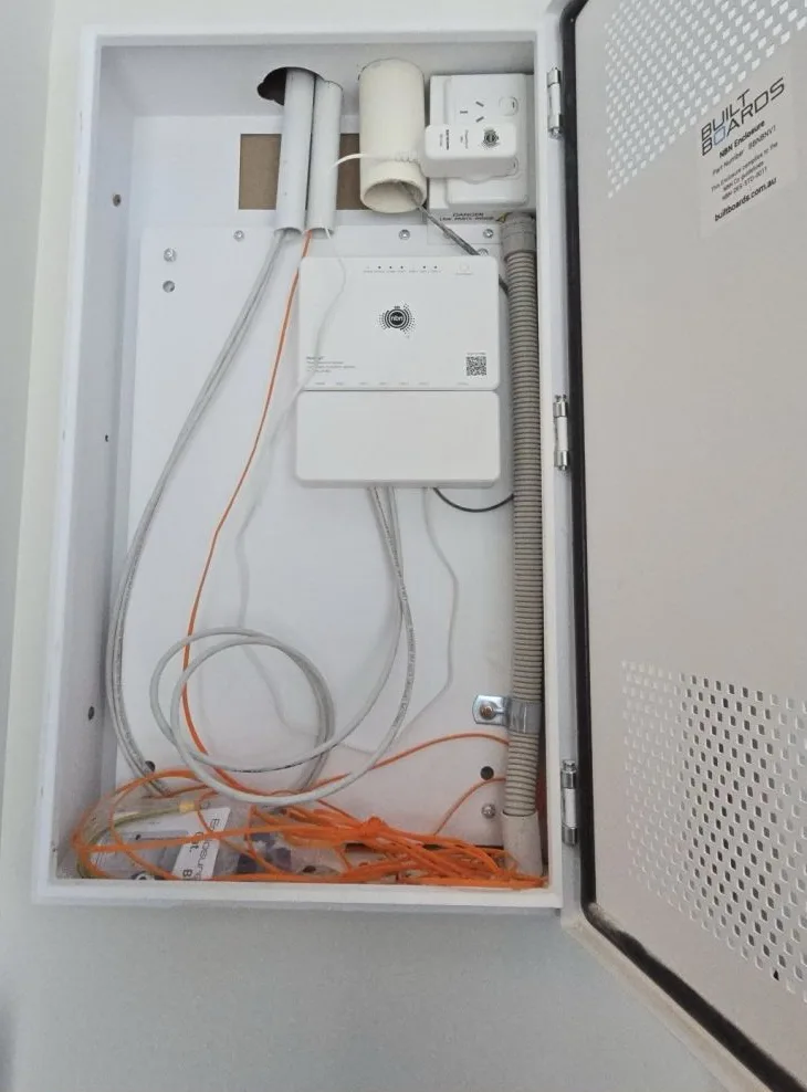 Open nbn enclosure after handover with orange leads knotted in the bottom, packaging left behind, no labels and conduit through a rough hole in the top.