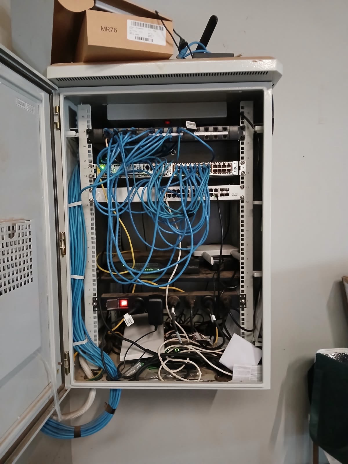 Wall-mounted network cabinet with an undocumented Meraki access point sitting on top and unmanaged patch cabling looped inside