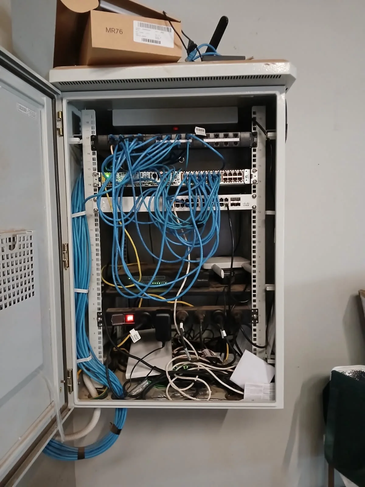 Wall-mounted network cabinet with an undocumented Meraki access point sitting on top and unmanaged patch cabling looped inside
