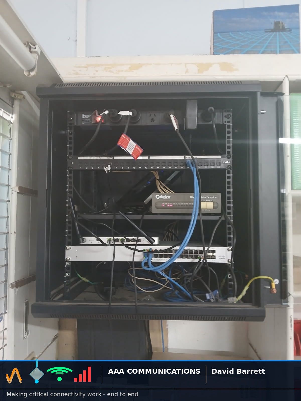 Wall-mounted network cabinet with a Telstra NTU, Cisco switch and patch panel, tangled patch leads below and a framed print resting on top